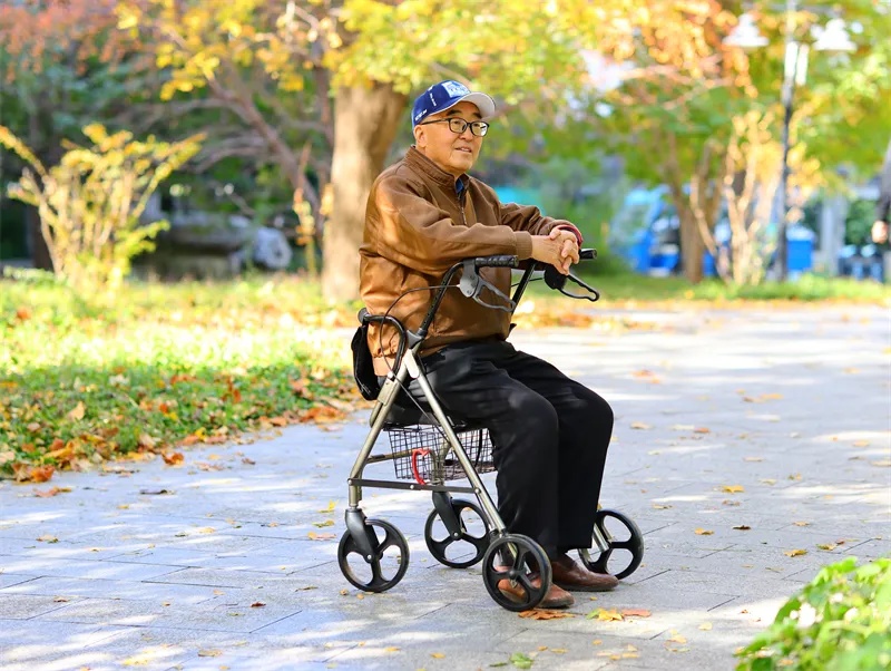 Elderly person using a Rollator with seat in a park setting, resting comfortably under a tree.