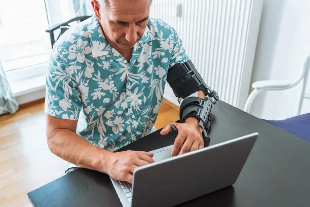 Illustration of the Wrist Carpal Tunnel Splint in use on a person's wrist while typing on a laptop. The scene is in a modern home office setting. The splint is clearly visible, providing support without hindering finger movement on the keyboard.