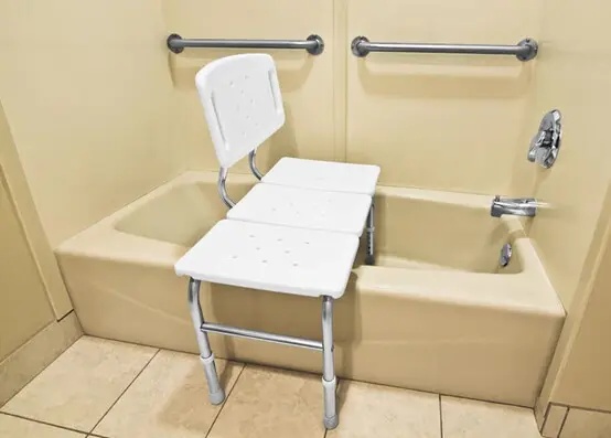 Shower Chair Commode in a clinical or care facility setting: The chair positioned next to a hospital bed, showing its role in patient transfer and mobility within an institutional context.