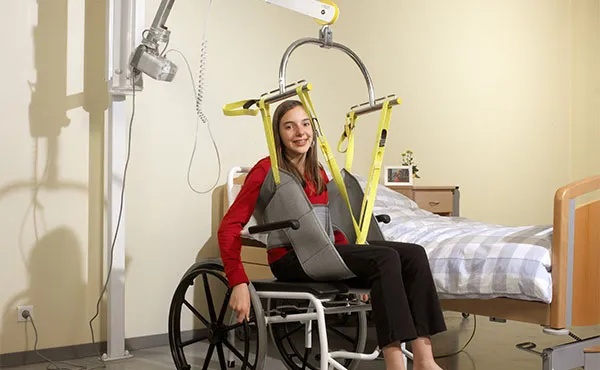 Hospital room scene showing a Manual Or Electric Lift Transfer Chair being used by a nurse to transfer a post-operative patient from the hospital bed to a wheelchair. The environment is clean and clinical.